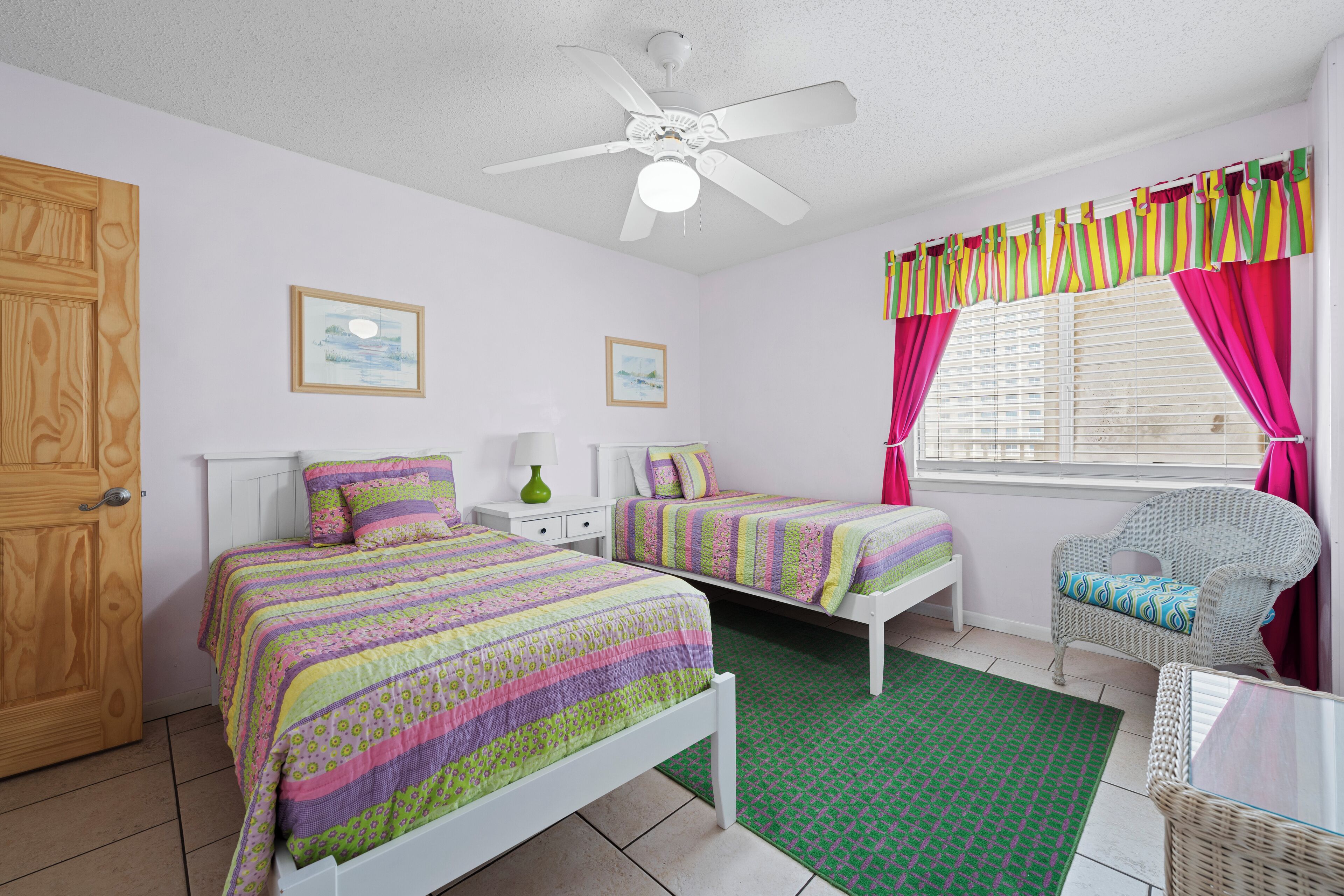 A bedroom with two beds, a ceiling fan, a window with curtains, and a wicker chair.