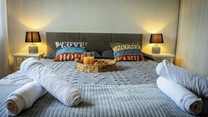 Un lit bien fait, recouvert d’une couverture grise à texture intéressante, avec deux oreillers ornés de motifs, un panier en osier contenant une bougie, et deux lampes de chevet posées sur des tables de nuit blanches.