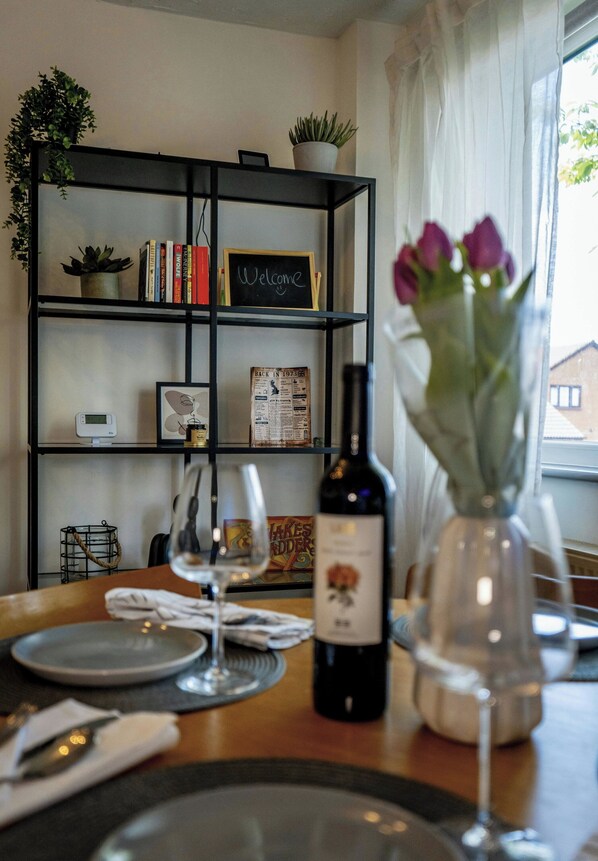 Un comedor con una mesa preparada para dos, una botella de vino, un jarrón con flores y una estantería con libros y un cartel de bienvenida.