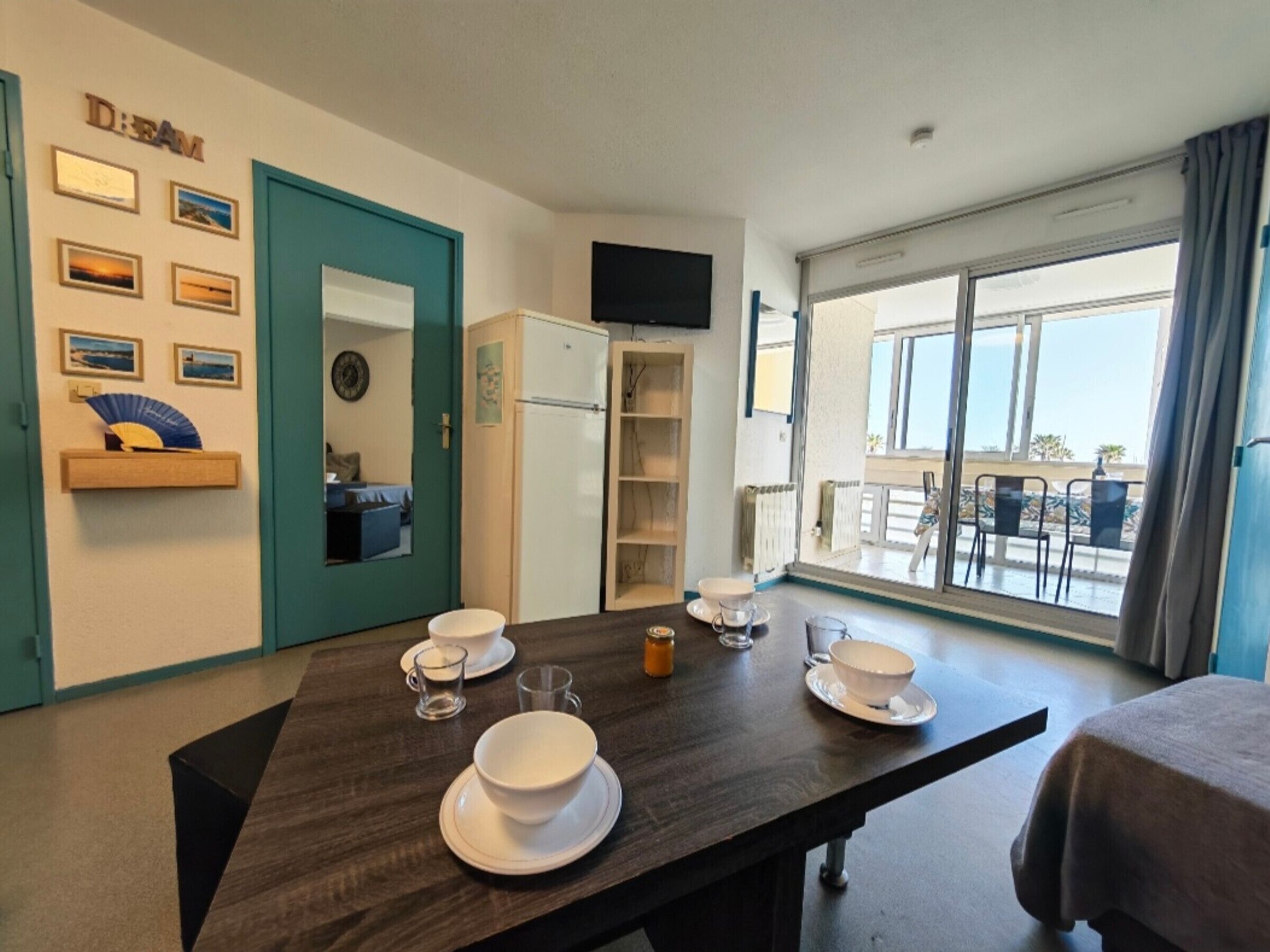 A hotel room with a dining table set with bowls and glasses, a TV mounted on the wall, a balcony with a view, and a teal-colored door.