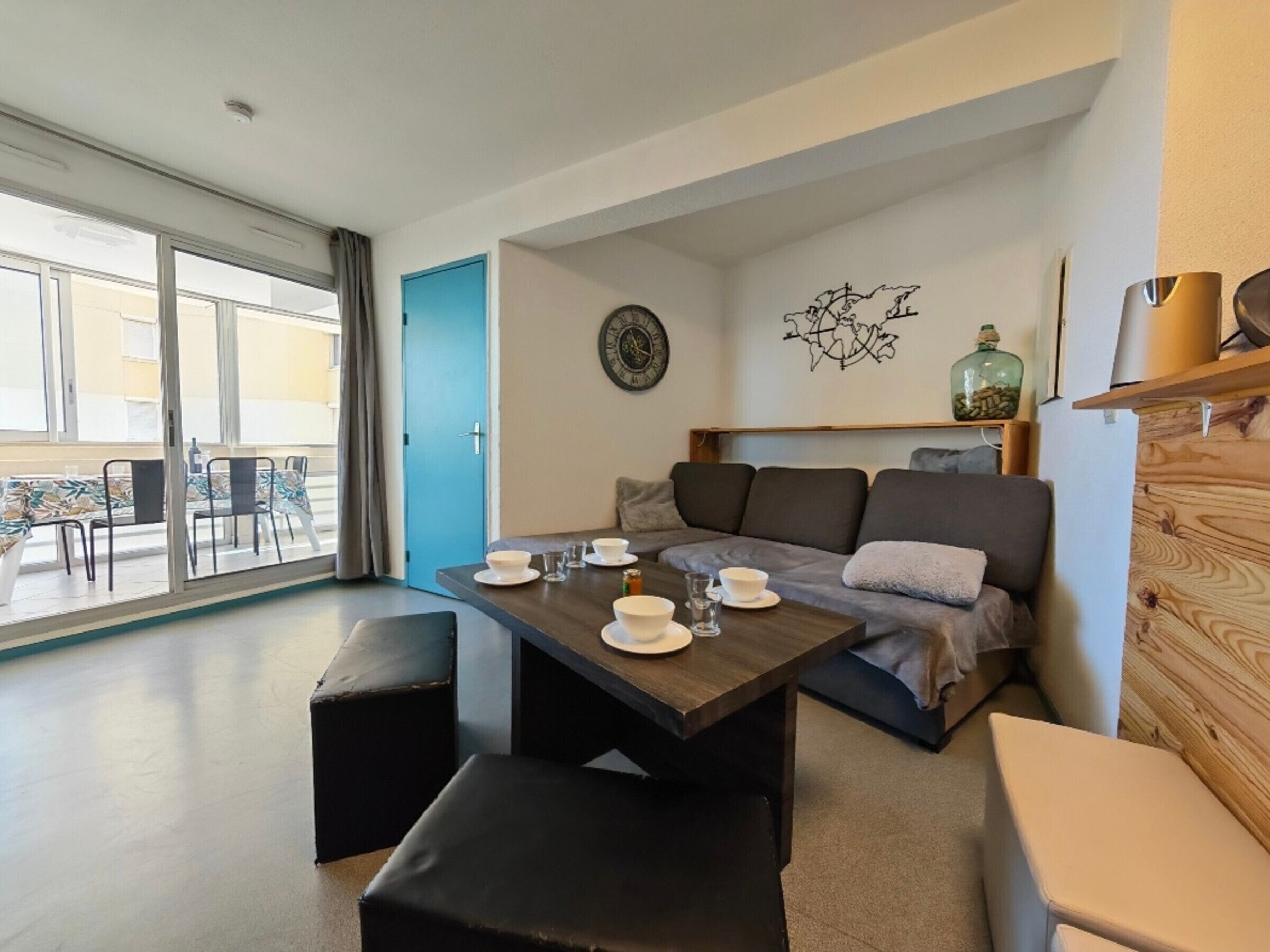 Un salon moderne avec un canapé, une table à manger dressée avec des bols et des verres blancs, une horloge et un élément décoratif mural.