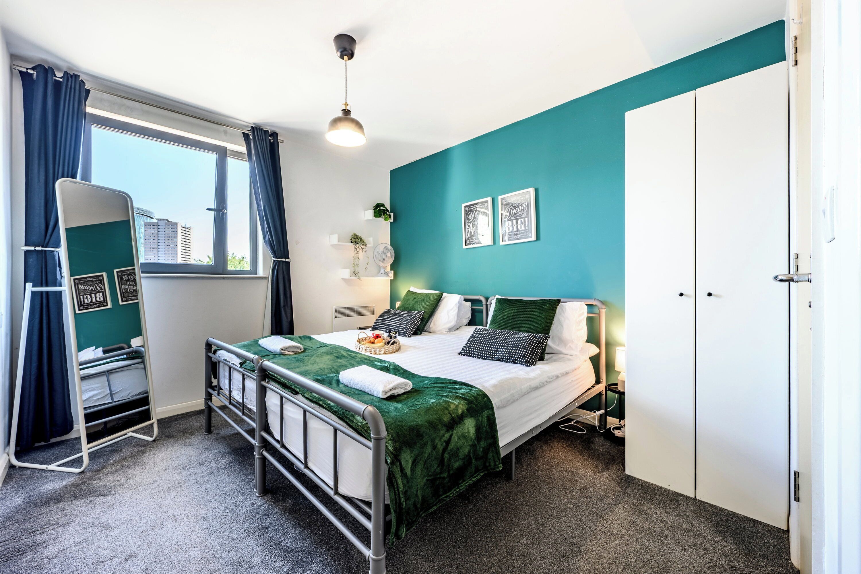 A bedroom with a bed, a bedside table, a mirror, a window with curtains, and a door.