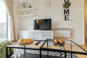 A living room with a TV, a basket of fruit, a plate with croissants, and a glass table.