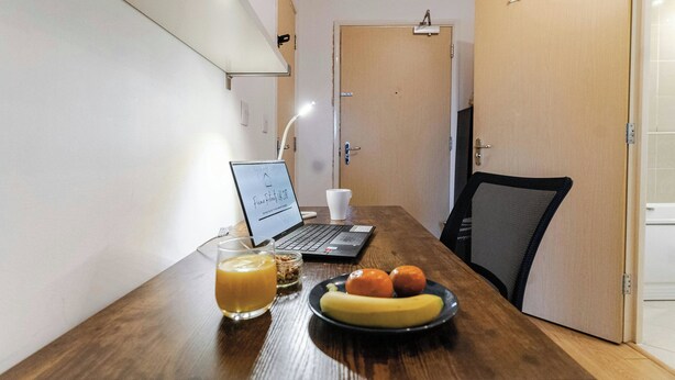 Et træbord med en bærbar computer, et glas appelsinjuice, en tallerken med en banan og to appelsiner, samt en sort mesh-stol.