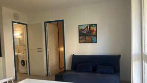 A living room with a blue sofa, a dining table, and a washing machine in the background. - Saint-Cyprien: Ground Floor T3, Terrace, Parking, 600m from Beach (Saint-Cyprien)