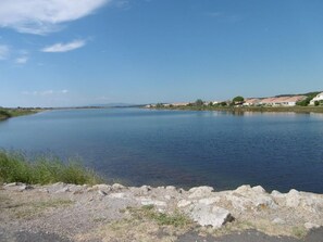 A calm lake with a rocky shoreline, surrounded by residential buildings and sparse vegetation. - Cozy T2 at Gruissan Beach: Pool, Parking, Terrace for 4, Near Shops (Gruissan)