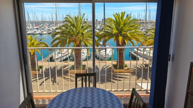 Un balcon avec une table et des chaises, donnant sur une marina où l’on peut admirer des bateaux et des palmiers.