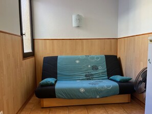 Un sofá de esquina con una manta color turquesa, paneles de madera en la pared, una lámpara pequeña fijada a la pared y un ventilador.