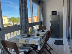 A balcony with a table set for two, a refrigerator, and a view of buildings and trees.
