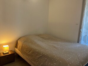 Une petite chambre avec un lit simple, un mur uni, une table de chevet avec une lampe et une fenêtre avec un rideau.