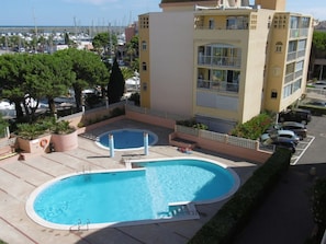 A swimming pool with a sunbathing area, surrounded by a building with balconies and a view of the marina.