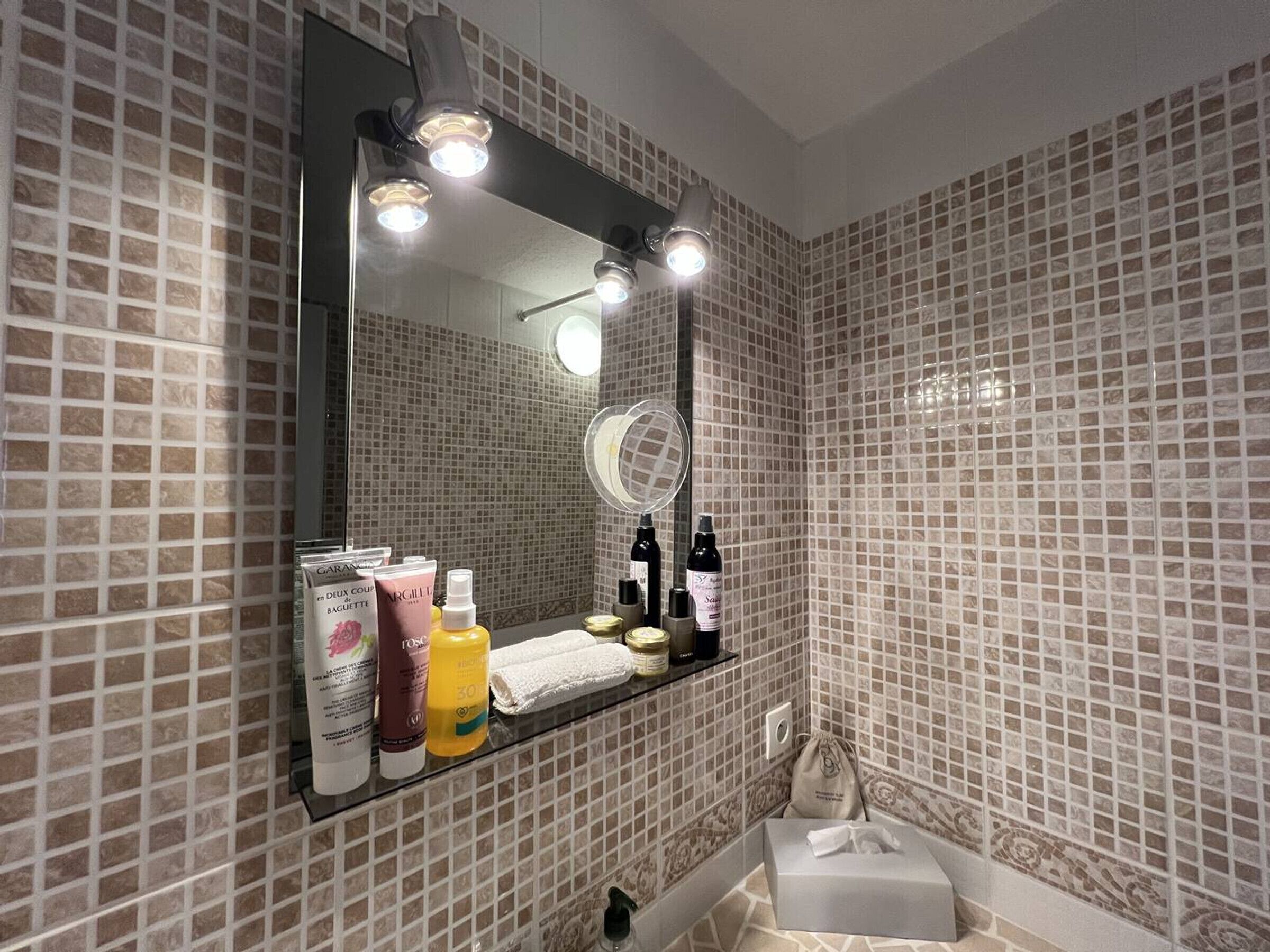 Une salle de bain avec un mur recouvert de carreaux derrière le lavabo, un miroir et des articles de toilette rangés sur une étagère.