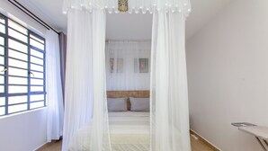 Un dormitorio con cama bajo un dosel, una ventana con persianas y una plancha pequeña.