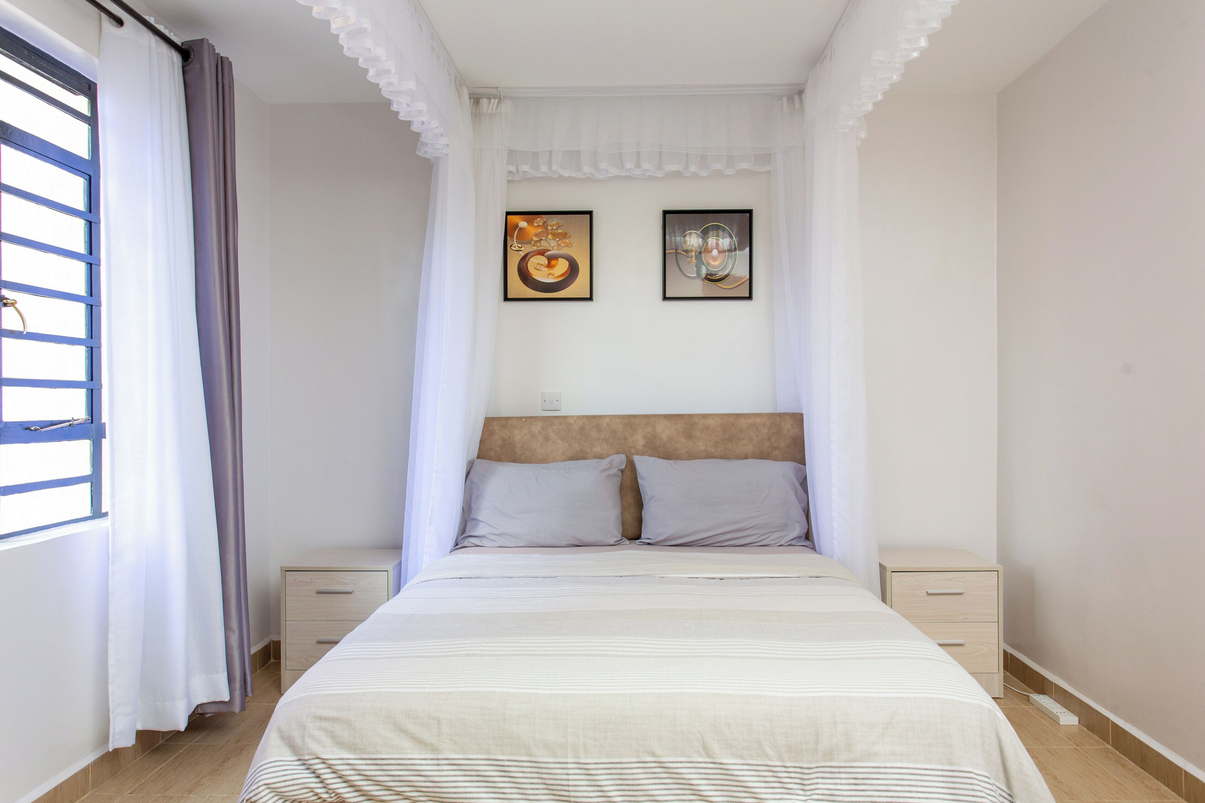 Un dormitorio con cama, dos mesitas de noche, una ventana con cortinas y dos cuadros en la pared.