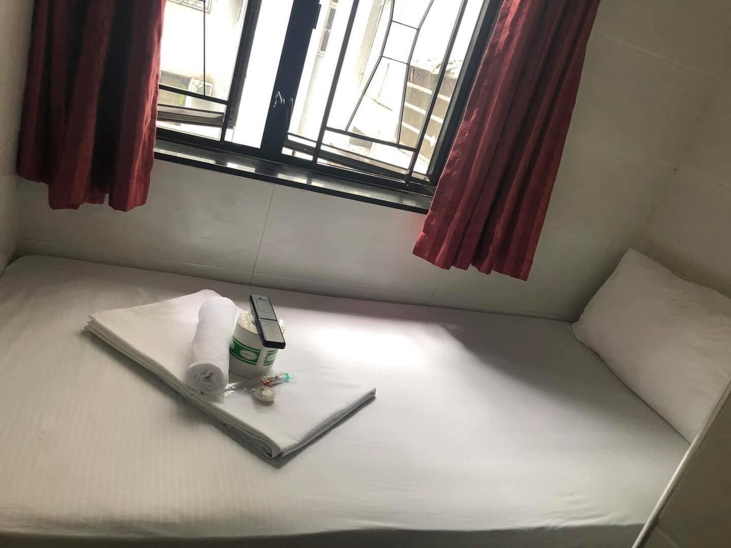 Ein kleines Zimmer mit einem Einzelbett, einem Fenster, einem Nachttisch mit einer Flasche und einer Taschentuchbox, sowie roten Vorhängen.