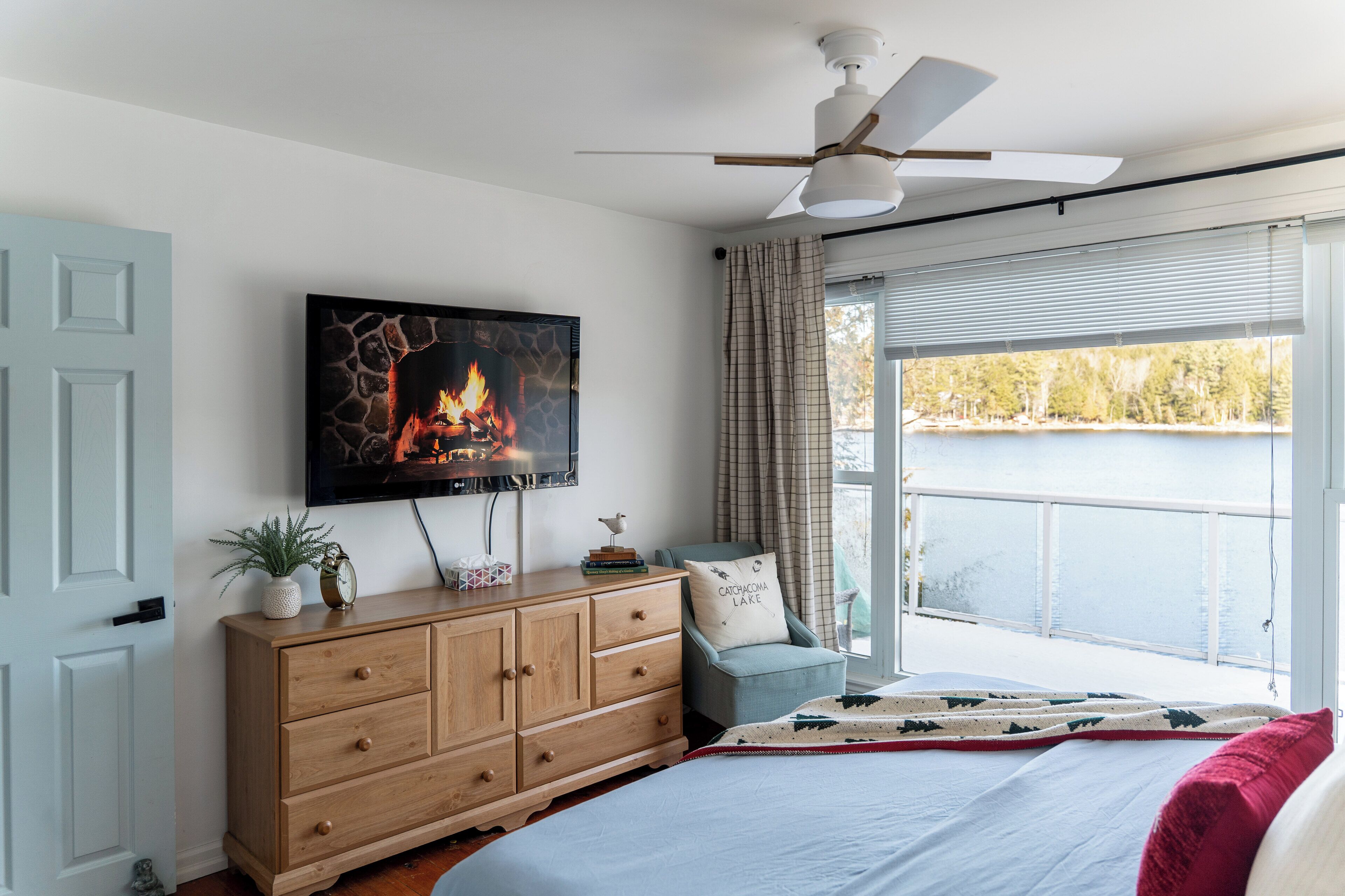 Une chambre avec un lit, une commode en bois, un téléviseur fixé au-dessus, et une vue sur un lac, visible à travers une porte-fenêtre coulissante.