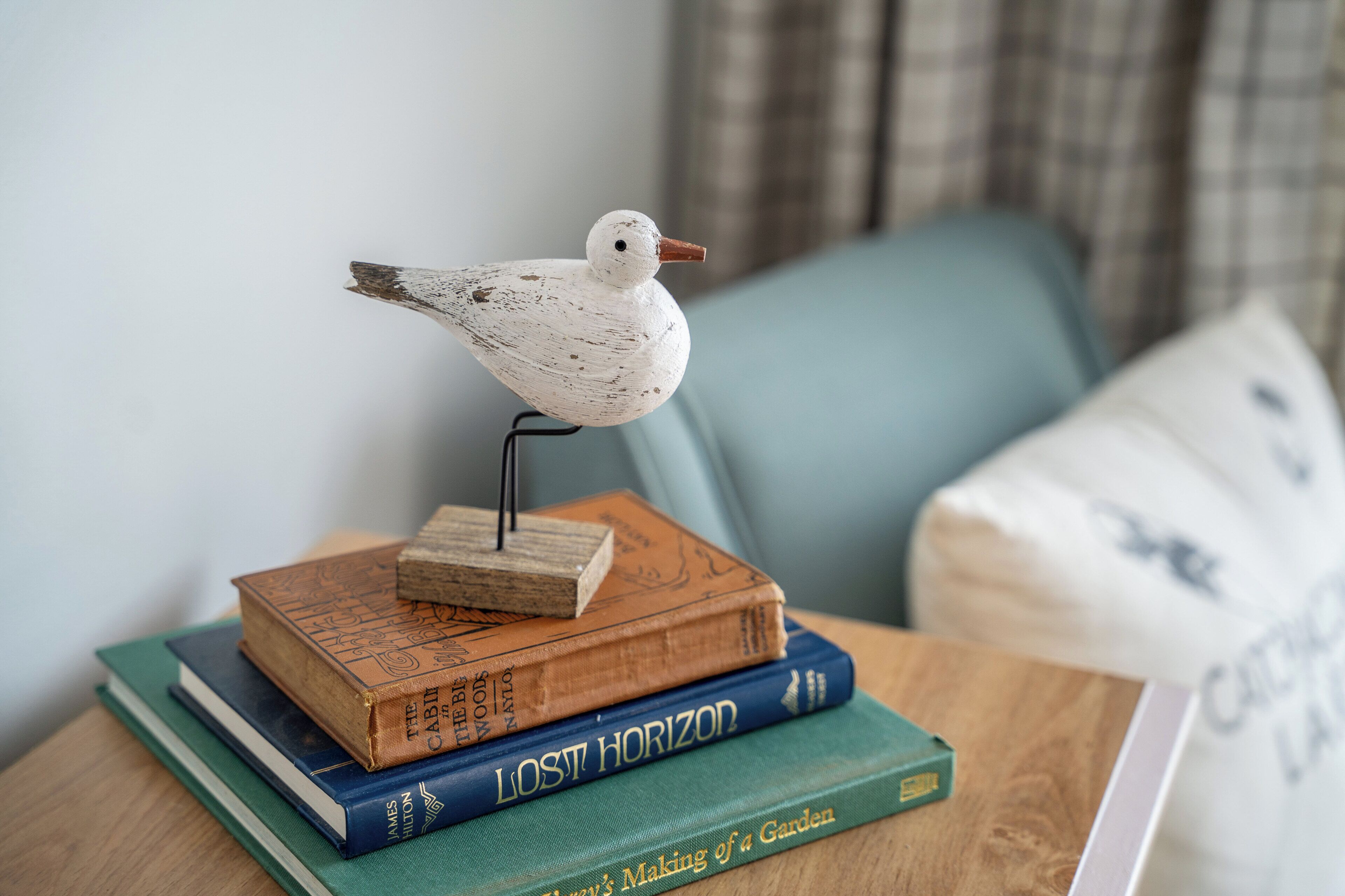 Ein Holztisch mit einem dekorativen Vogel, mehreren Büchern und einem Kissen im Hintergrund.