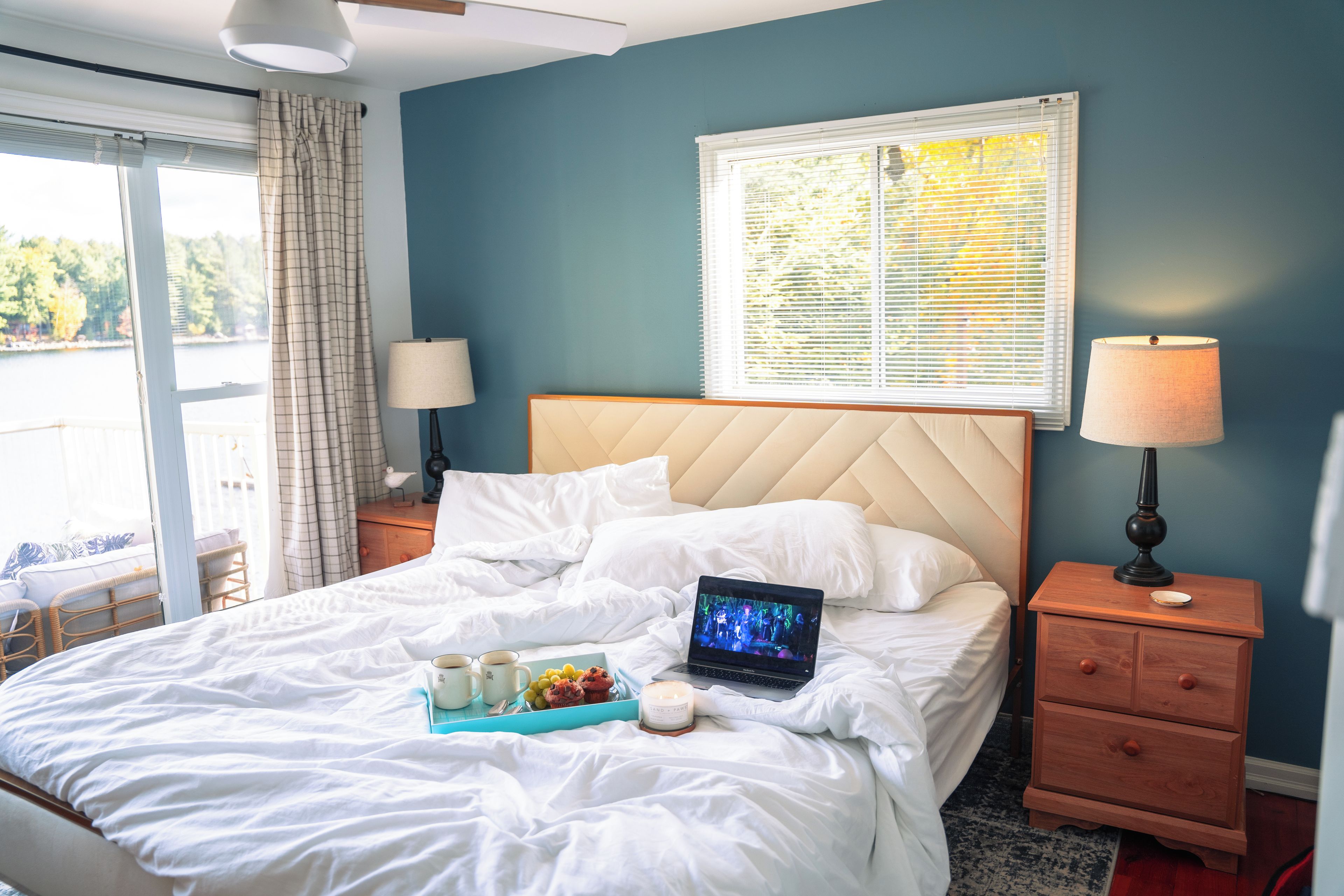Una cama bien hecha con una computadora portátil, una bandeja con comida y bebidas, y una ventana con persianas.