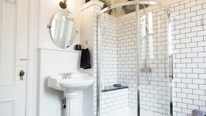 Une salle de bain avec un lavabo sur pied blanc, un miroir rond et une douche à parois de verre, les murs étant recouverts de carreaux blancs.