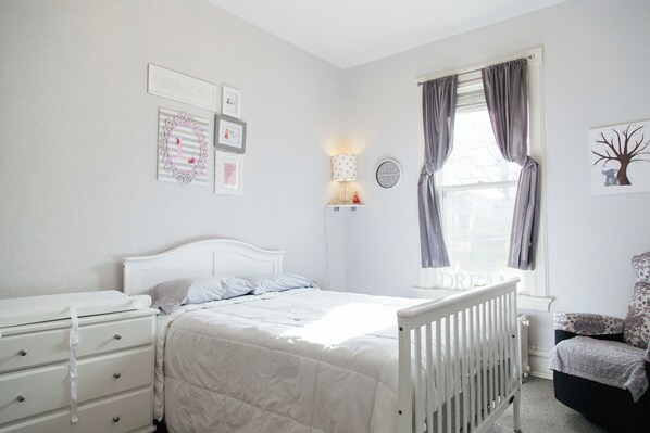 Un dormitorio con cama blanca, cuna, cómoda, mesita de noche, lámpara y ventana con cortinas.
