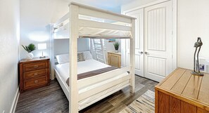 A bunk bed with a wooden frame and white bedding, a wooden dresser, a nightstand with a lamp, and a door with a white panel. - Beachside Heated Pool/Hot Tub  2 King Beds  Firepit + Lift (Galveston)