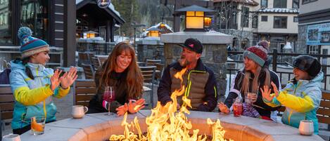 A group of people seated around a fire pit in an outdoor setting with buildings in the background.