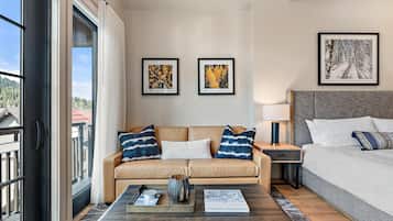 A modern hotel room with a sofa, a bed, a nightstand, and a coffee table. There are two framed pictures on the wall, a window with curtains, and a view of buildings outside.