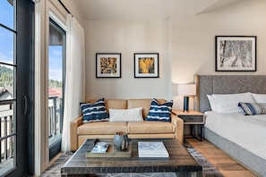 Habitación de hotel moderna con sofá, cama, mesita de noche y mesa de centro. Se observan dos cuadros en la pared, una ventana con cortinas y vistas a edificios.