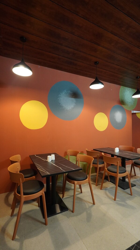 A restaurant interior with wooden ceiling beams, round tables with chairs, and wall art. - Hotel Hive Inn (Kozhikode)