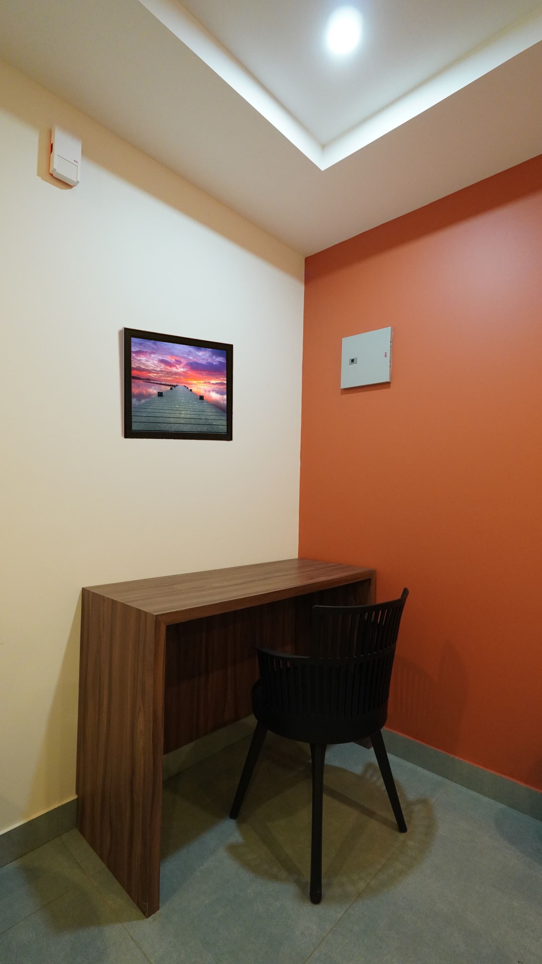 Une pièce avec un bureau en bois, une chaise noire, un tableau encadré au mur et un luminaire au plafond.