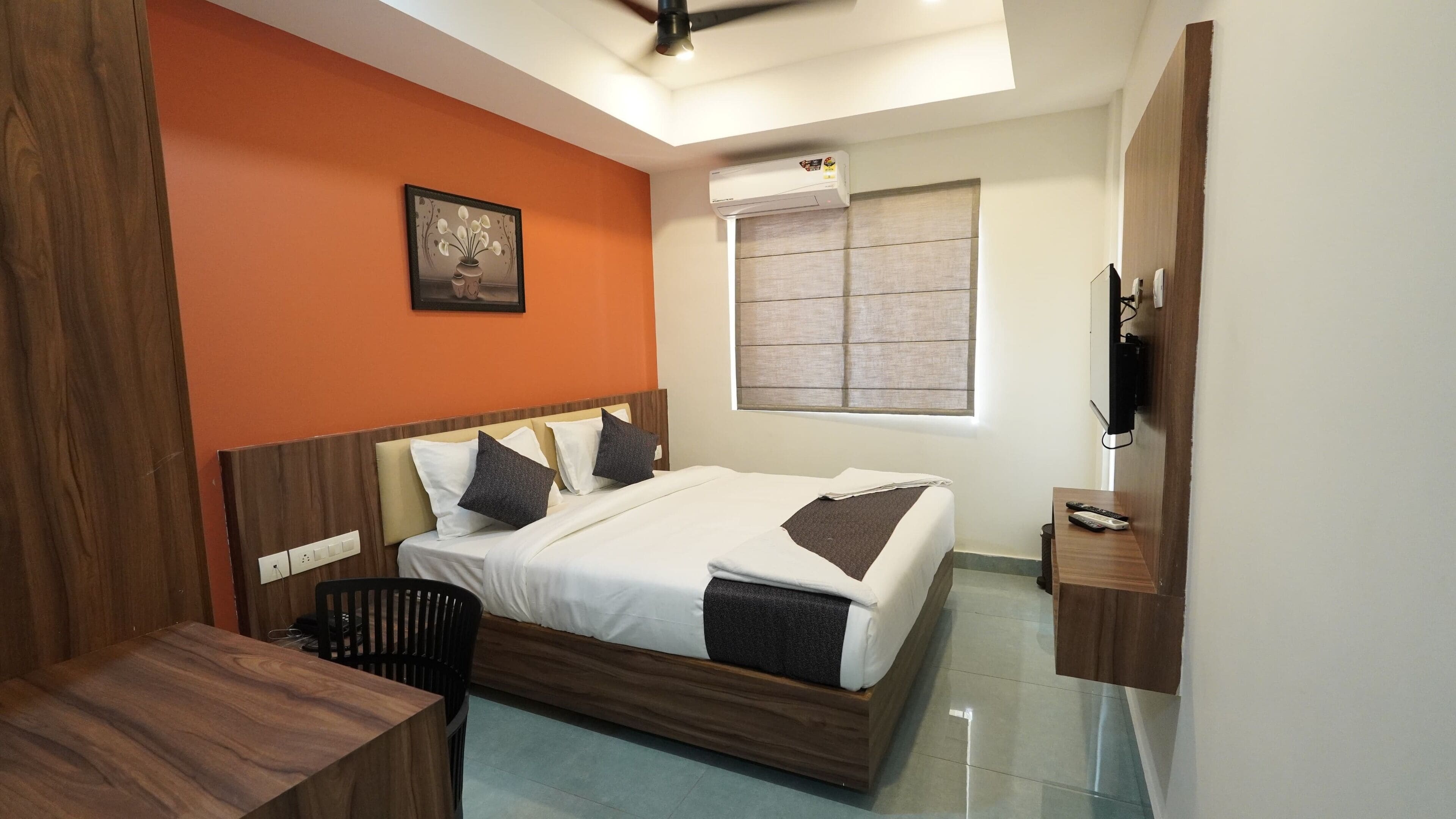 A hotel room with a bed, a desk, a chair, a TV, and an air conditioning unit.