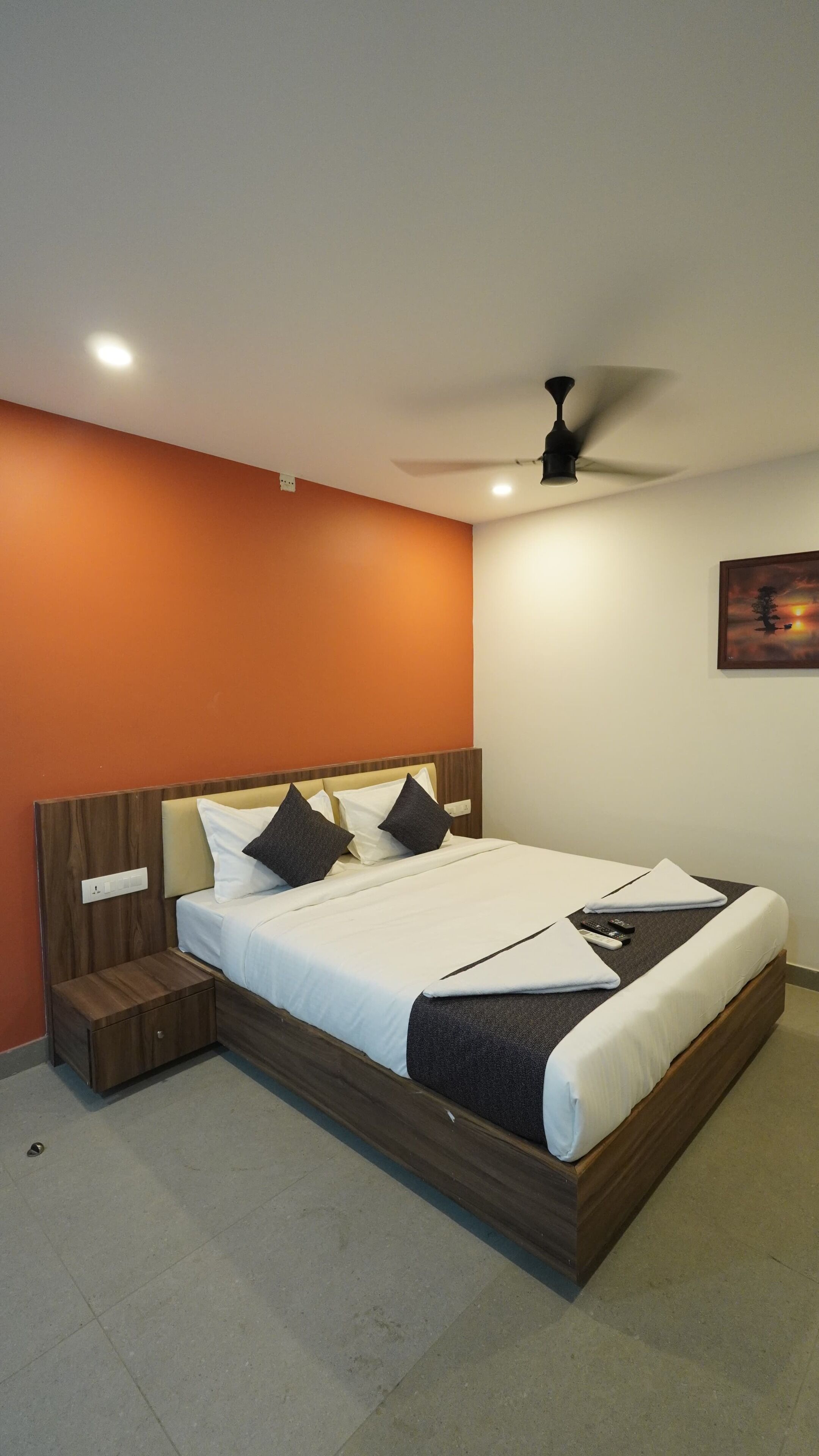 Uma cama bem arrumada com lençóis brancos e almofadas escuras, um criado-mudo de madeira, um ventilador de teto e um quadro na parede.