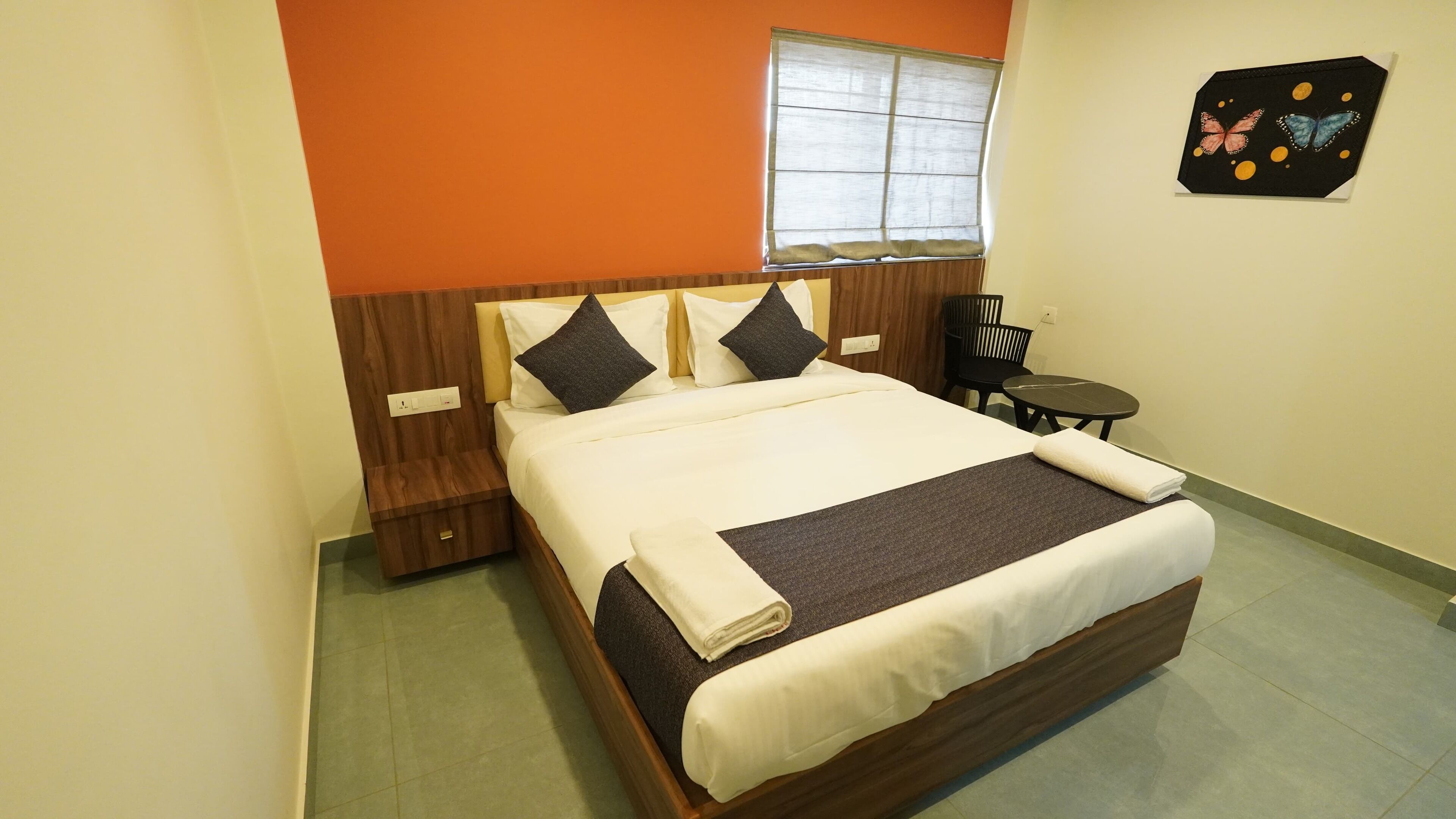 A neatly made bed with a white and brown bedspread, a wooden headboard, and two bedside tables with lamps. There is a window with blinds, a small round table, and a chair in the room.