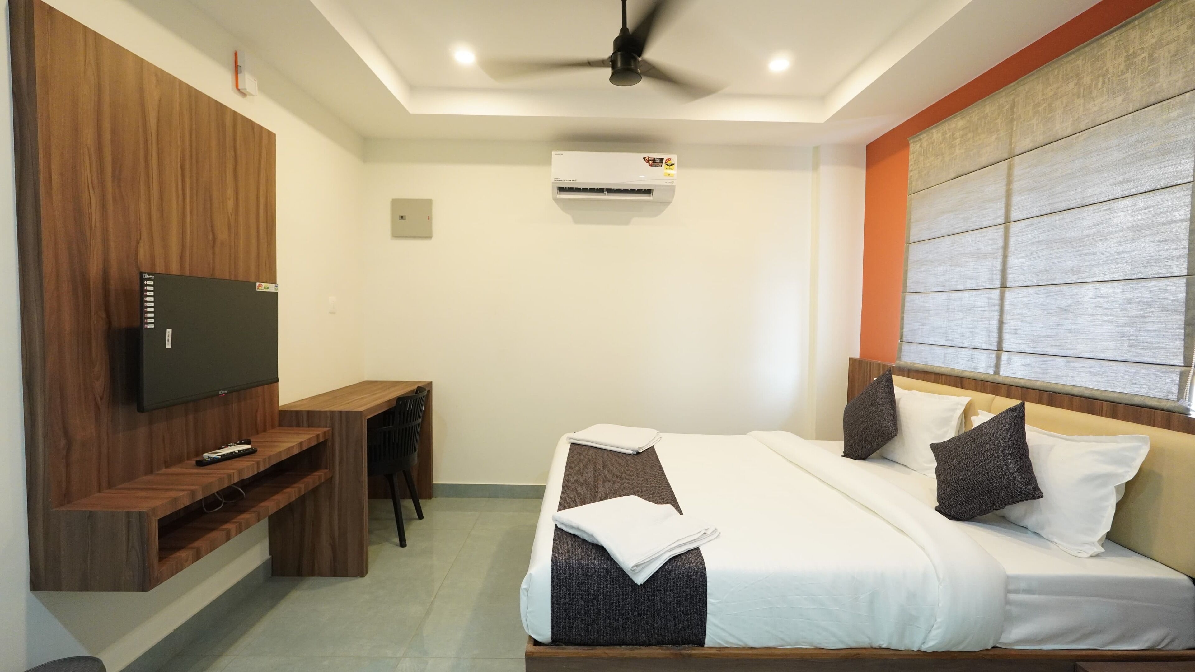 A hotel room with a bed, a TV mounted on a wooden panel, a desk with a chair, an air conditioning unit, and a ceiling fan.