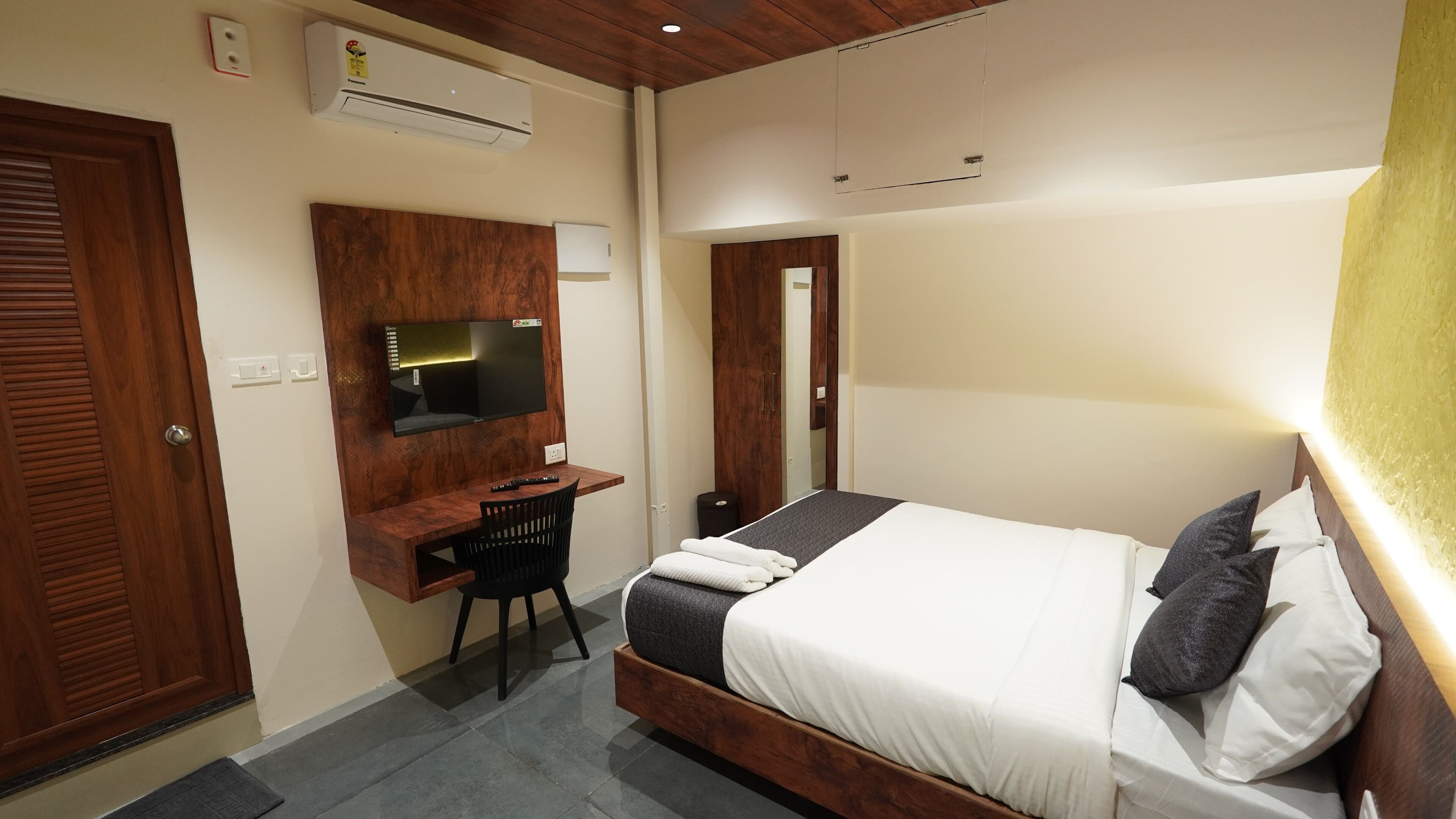 A hotel room with a wooden bed, a desk with a chair, a flat-screen TV, and a mirror.