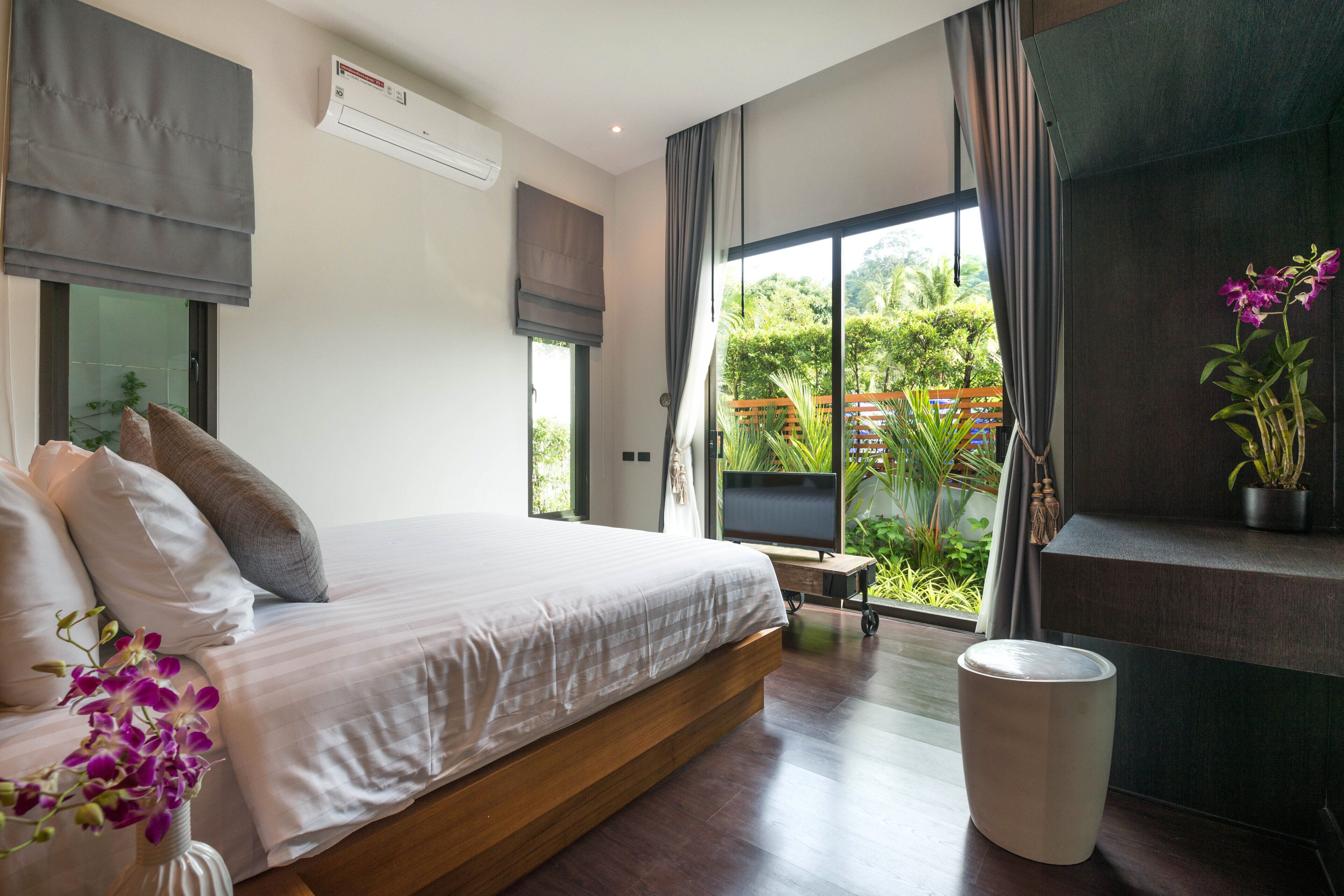 A bedroom with a bed, a TV, a window with curtains, a potted plant, and a waste bin.