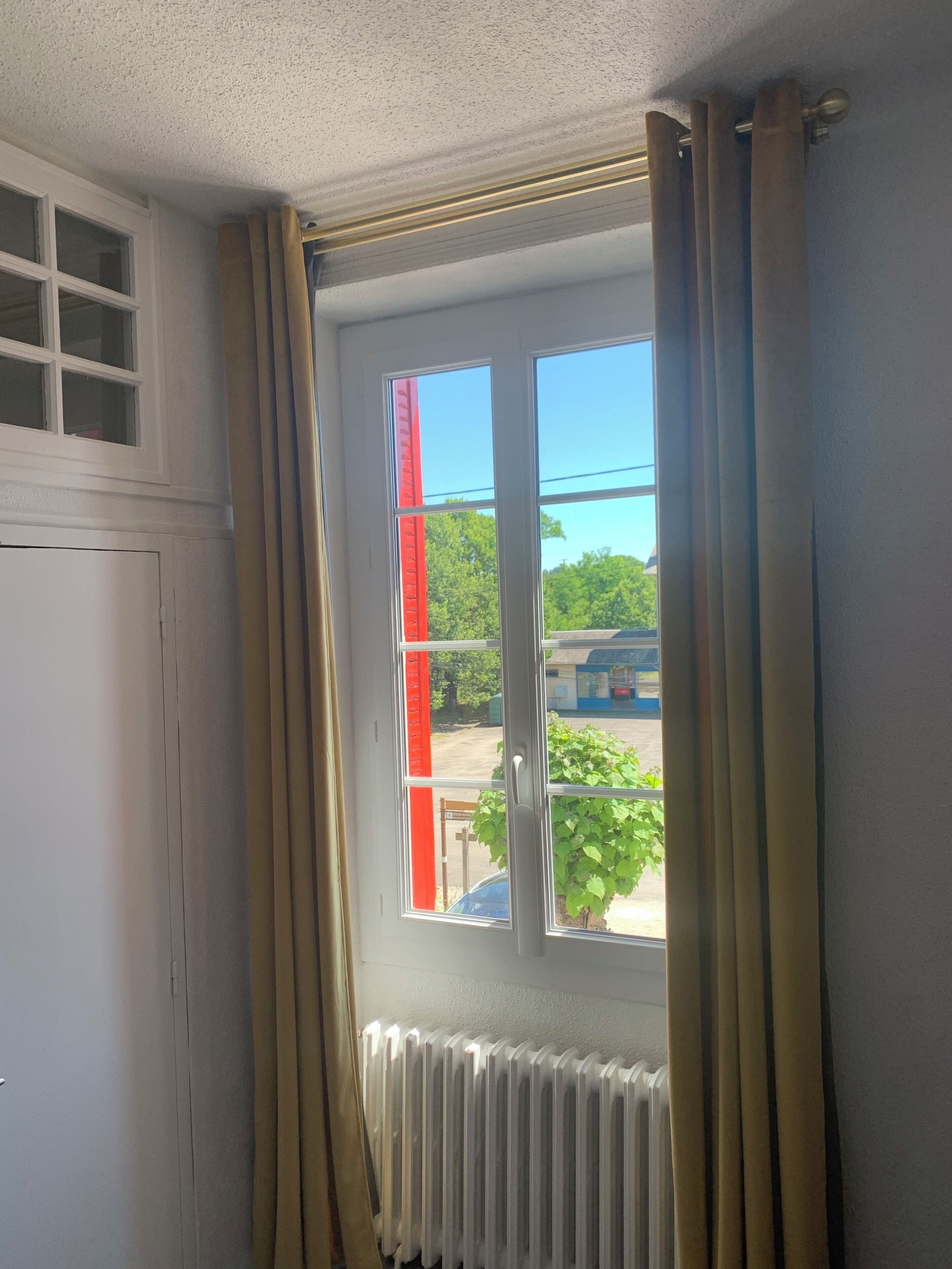 A room with a window, curtains, a radiator, and a view of a red structure outside.
