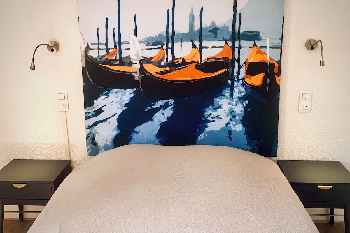 Een hotelkamer met een groot schilderij van boten aan de muur, een bed, twee nachtkastjes en wandlampen.