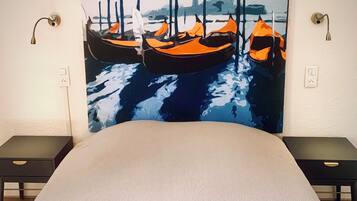 Habitación de hotel con una pintura grande de barcos en la pared, una cama, dos mesitas de noche y lámparas fijadas a la pared.