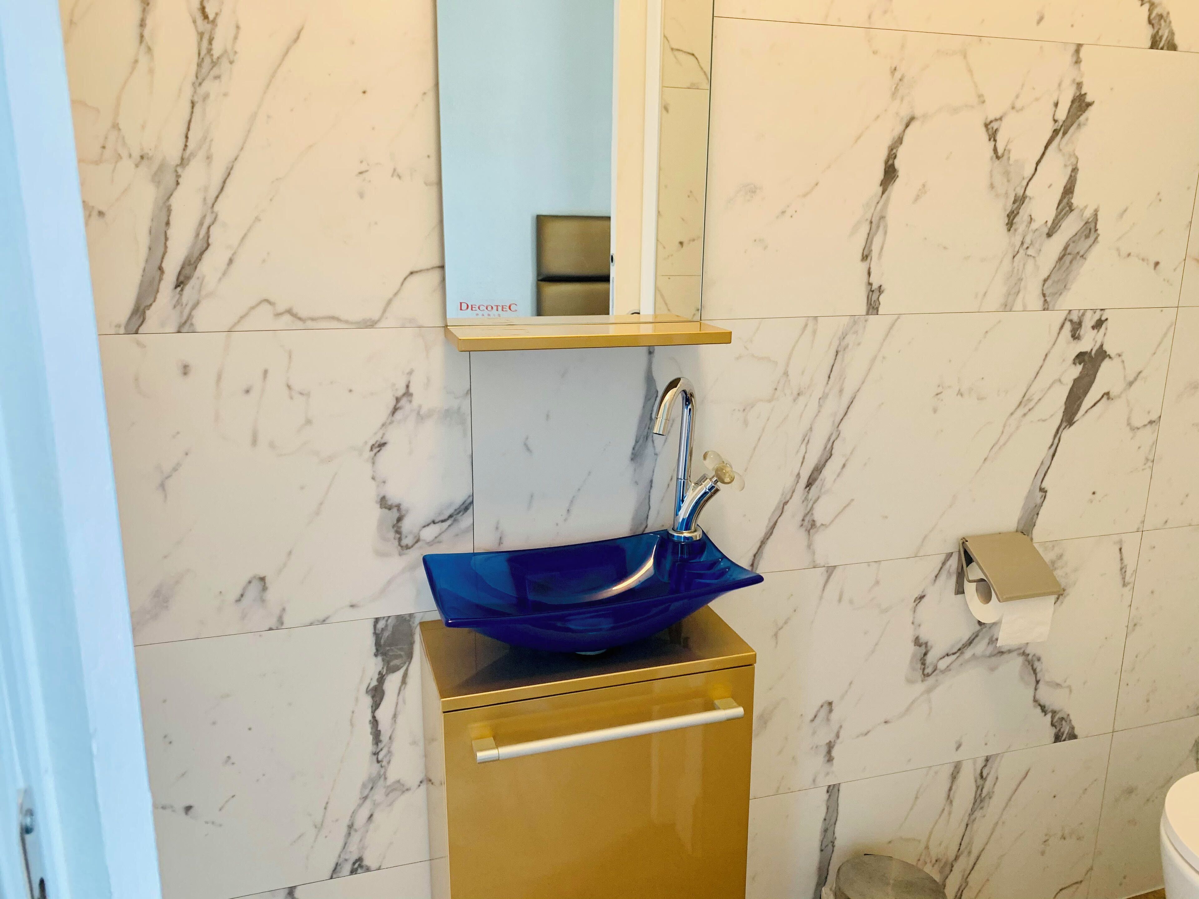 Un baño con pared de mármol, un mueble dorado, un lavamanos azul y un dispensador de papel higiénico.