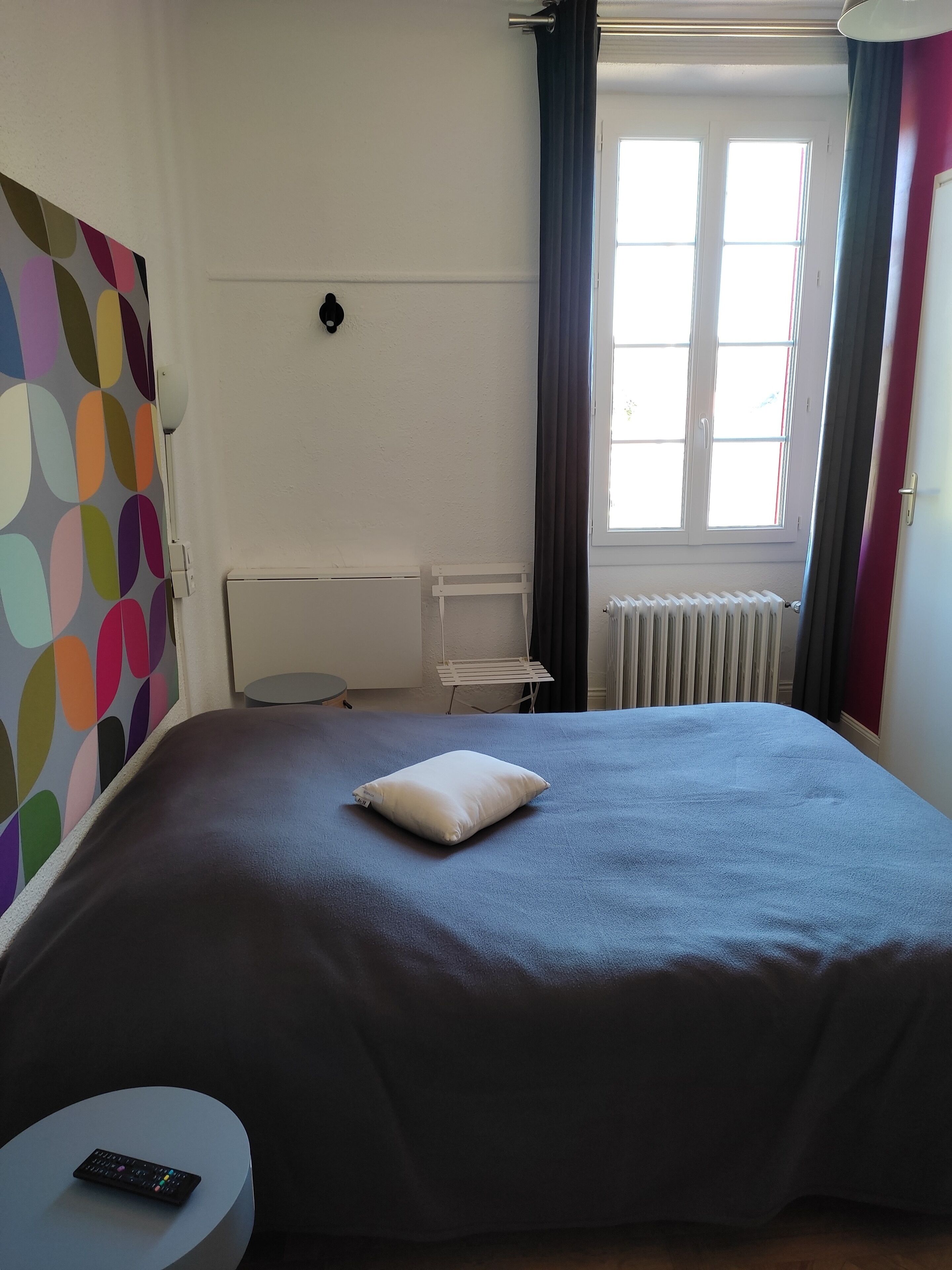 Un dormitorio con cama, una mesita redonda, una lámpara de pared, un radiador y una ventana con cortinas.