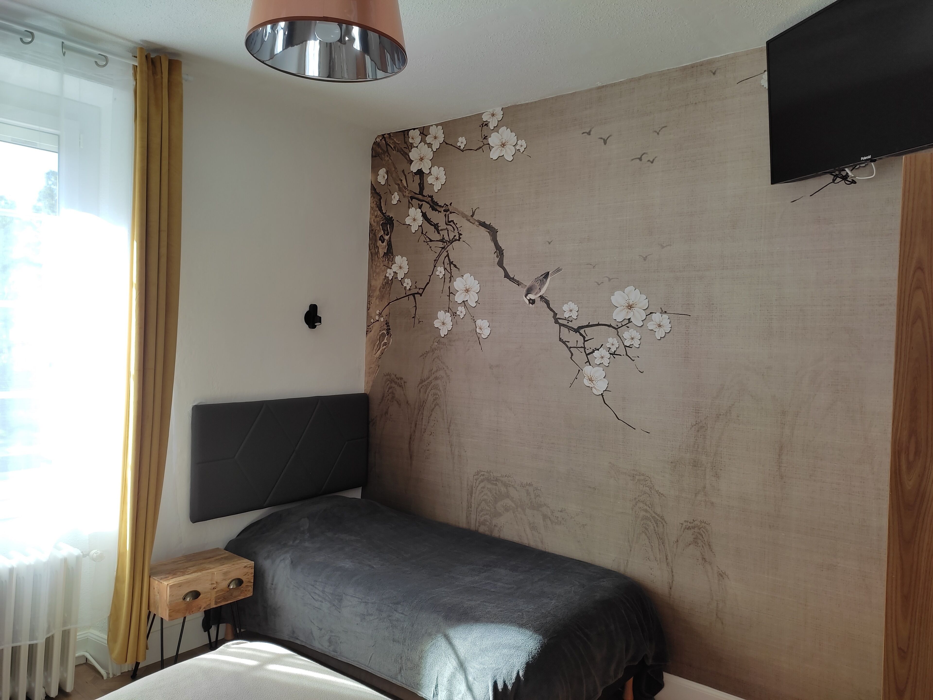 A bedroom with a bed, a bedside table, a wall-mounted TV, and a window with curtains.