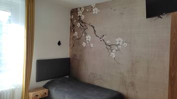 A bedroom with a bed, a bedside table, a wall-mounted TV, and a window with curtains.