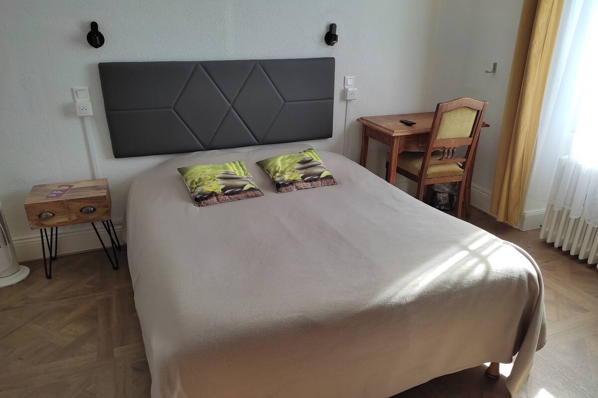 Een net opgemaakt bed met een groot hoofdeinde, een nachtkastje met een lamp en een stoel met een tafeltje.