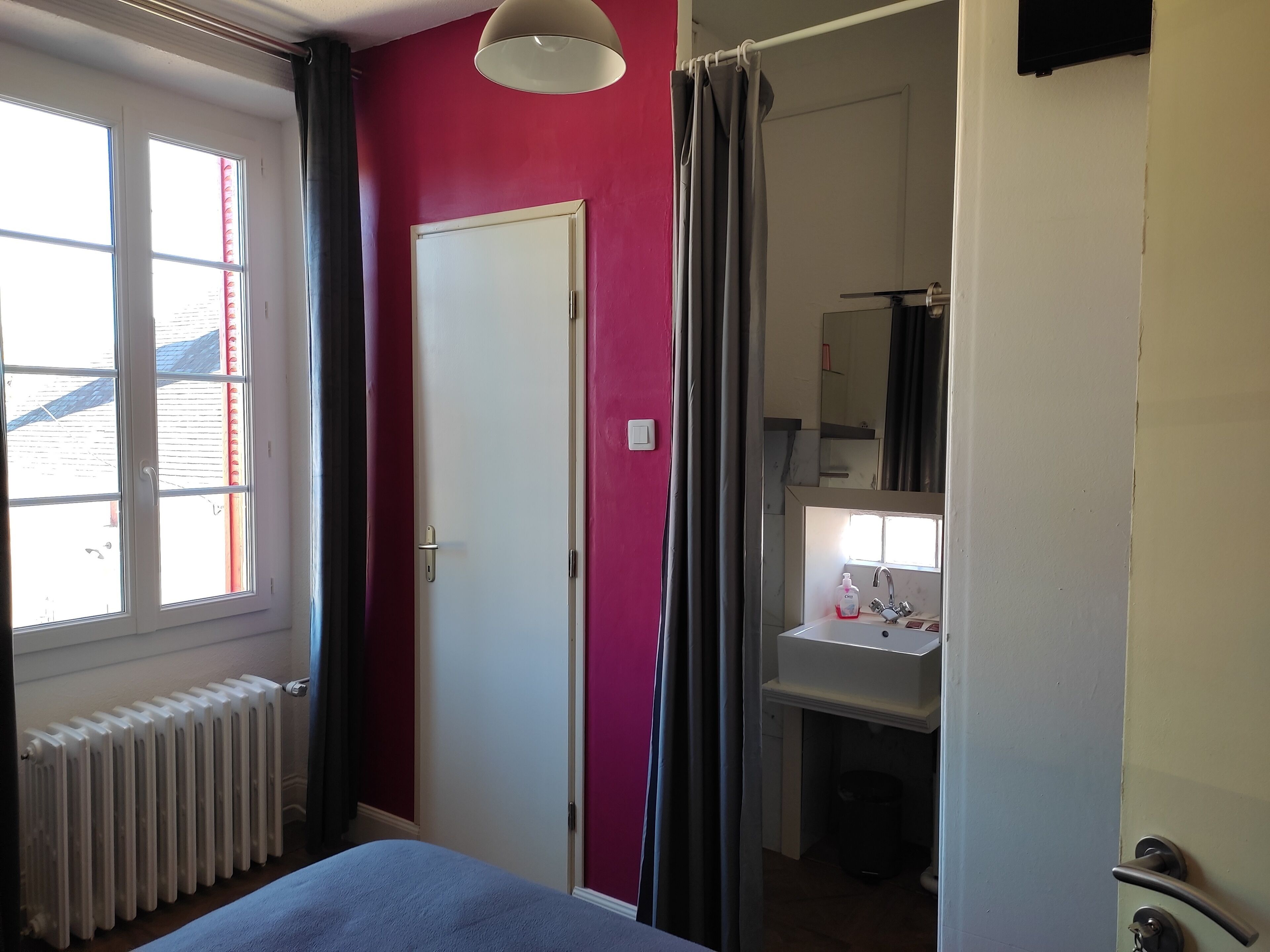 A room with a bed, a radiator, a window with curtains, a door, and a bathroom with a sink and mirror.