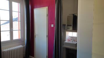 A room with a bed, a radiator, a window with curtains, a door, and a bathroom with a sink and mirror.
