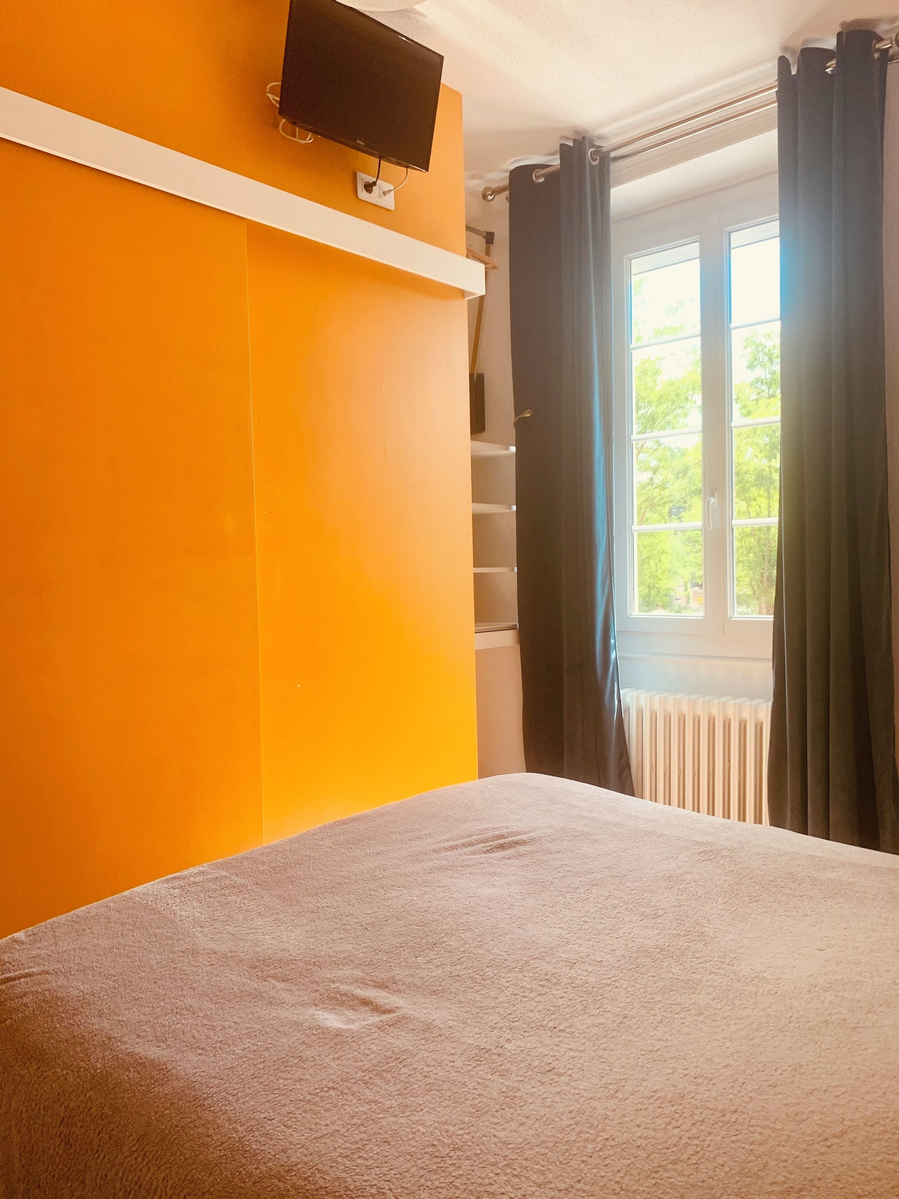 A bedroom with an orange wall, a bed, a TV mounted on the wall, and a window with curtains.