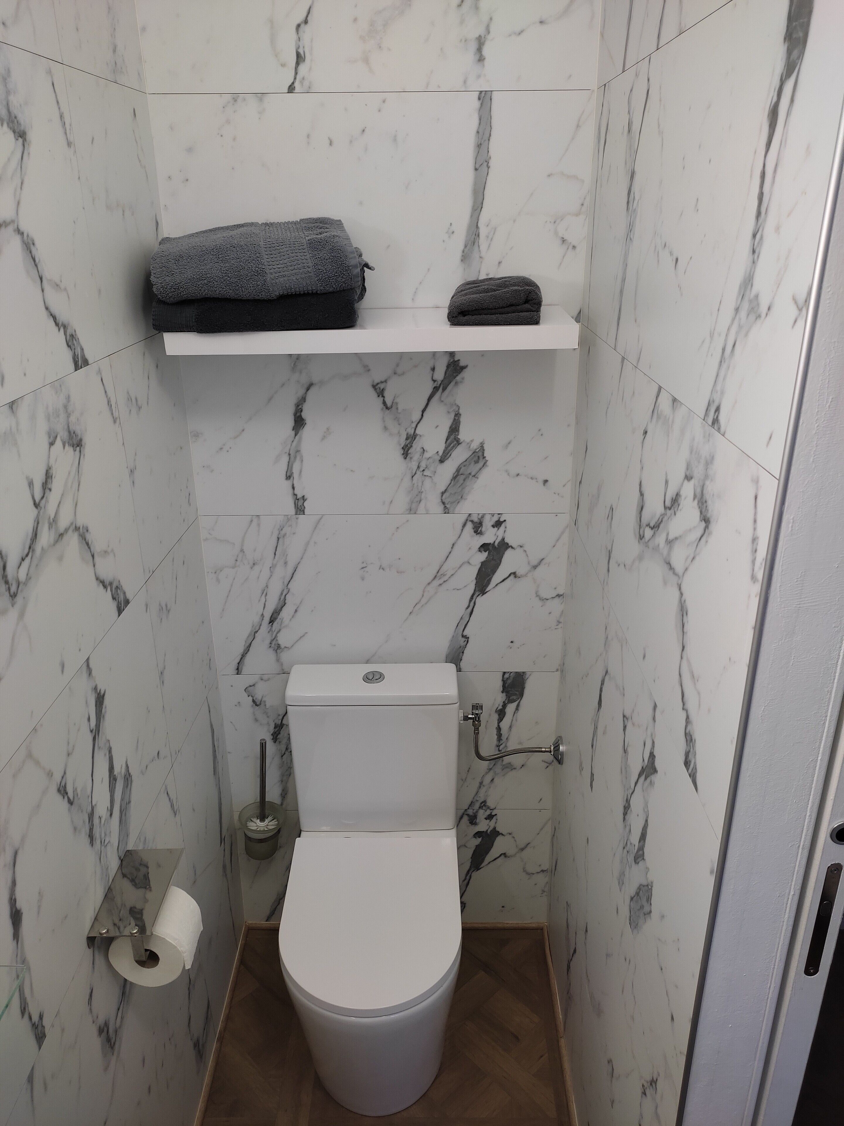 Un baño con inodoro, paredes con diseño de mármol, repisas con toallas y un dispensador de papel higiénico.