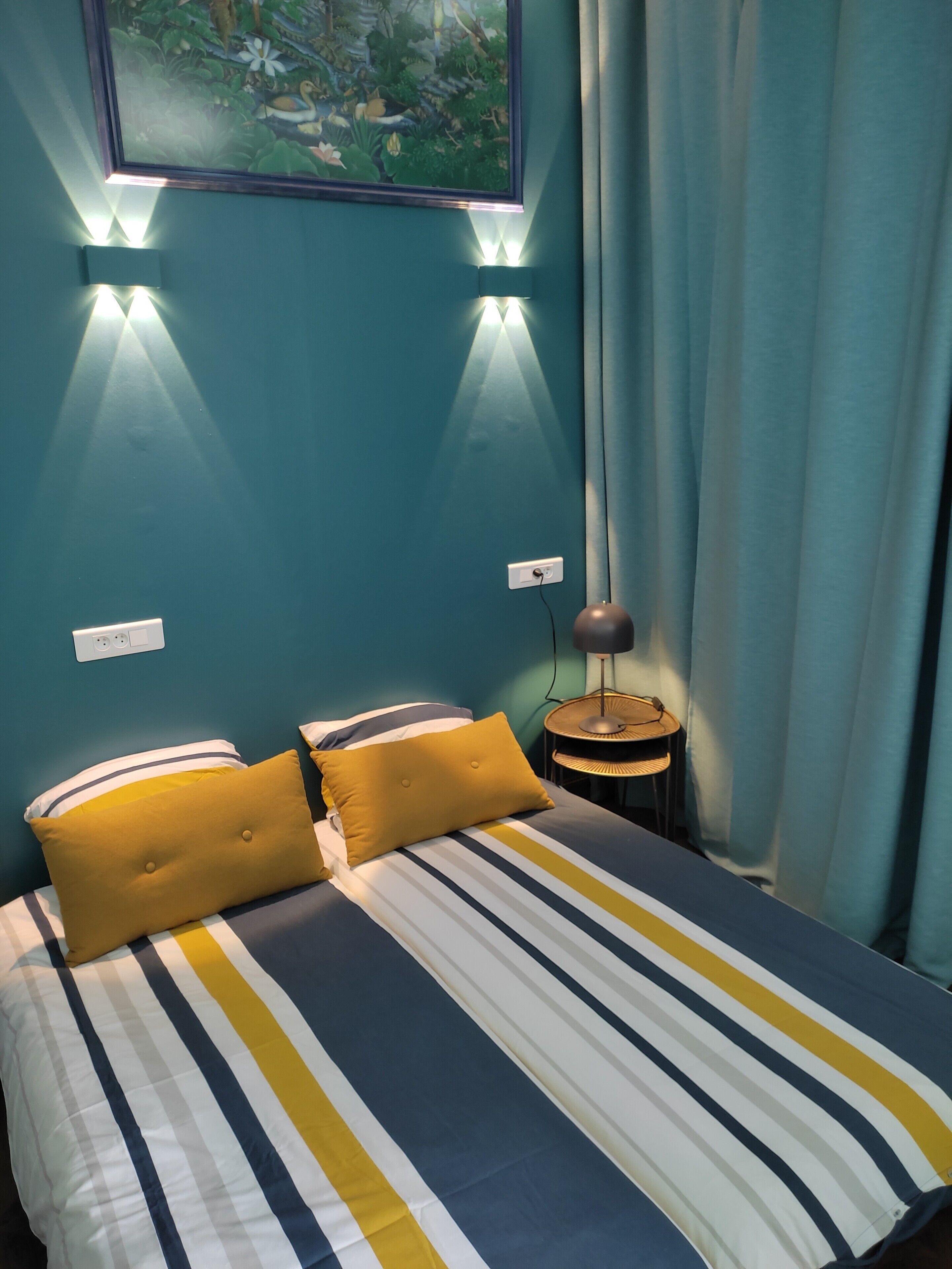 A bed with striped bedding and two mustard pillows, a small round table with a lamp, and a wall-mounted light fixture.