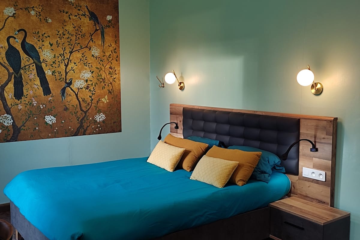 Een slaapkamer met een groot bed, twee nachtkastjes, een wandlamp en een schilderij van vogels in een boom.